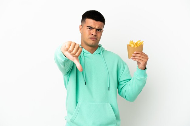 Hombre guapo joven sosteniendo patatas fritas sobre fondo blanco aislado mostrando el pulgar hacia abajo con expresión negativa