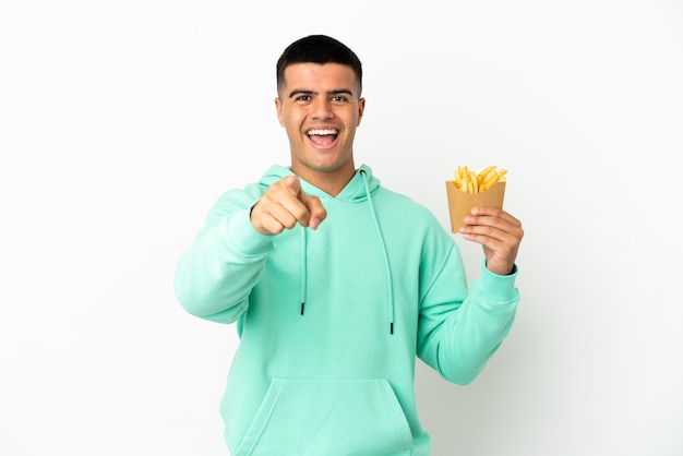 Hombre guapo joven sosteniendo patatas fritas sobre fondo blanco aislado sorprendido y apuntando hacia el frente