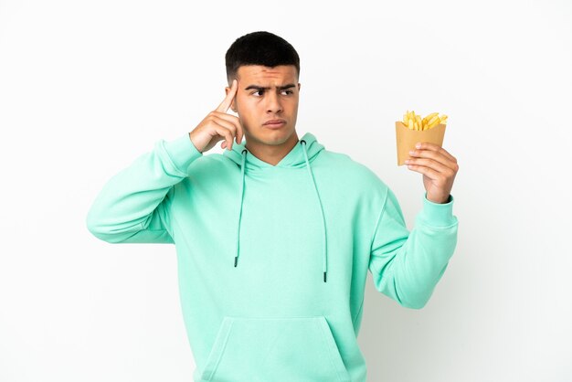 Hombre guapo joven sosteniendo patatas fritas sobre fondo blanco aislado teniendo dudas y pensando