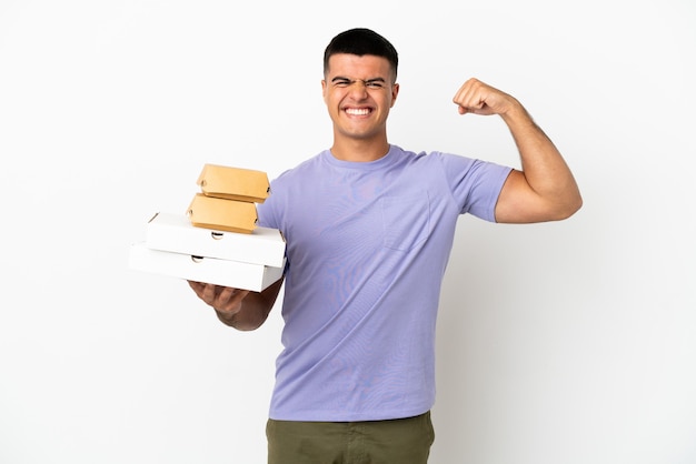 Hombre guapo joven sosteniendo pizzas y hamburguesas sobre fondo blanco aislado haciendo gesto fuerte