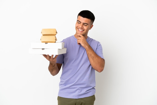 Hombre guapo joven sosteniendo pizzas y hamburguesas sobre fondo blanco aislado mirando hacia el lado y sonriendo