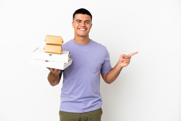 Hombre guapo joven sosteniendo pizzas y hamburguesas sobre fondo blanco aislado que señala con el dedo hacia el lado