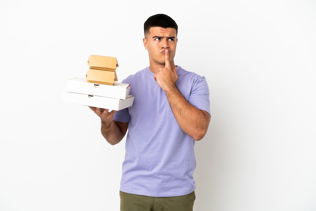 Hombre guapo joven sosteniendo pizzas y hamburguesas sobre fondo blanco aislado que tiene dudas mientras mira hacia arriba