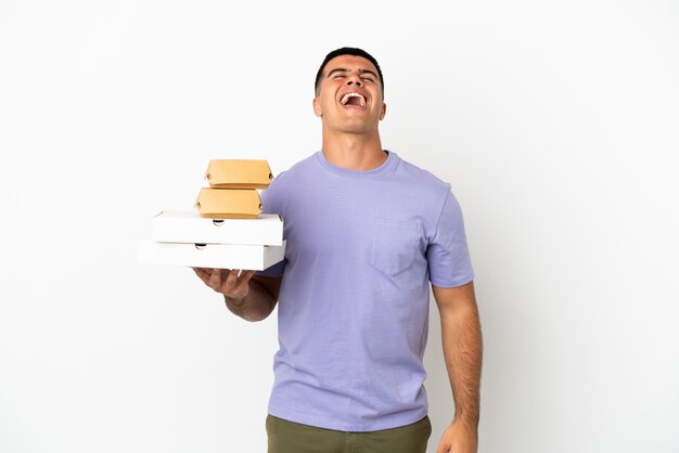 Hombre guapo joven sosteniendo pizzas y hamburguesas sobre fondo blanco aislado riendo