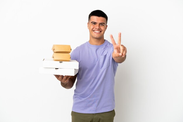 Hombre guapo joven sosteniendo pizzas y hamburguesas sobre fondo blanco aislado sonriendo y mostrando el signo de la victoria