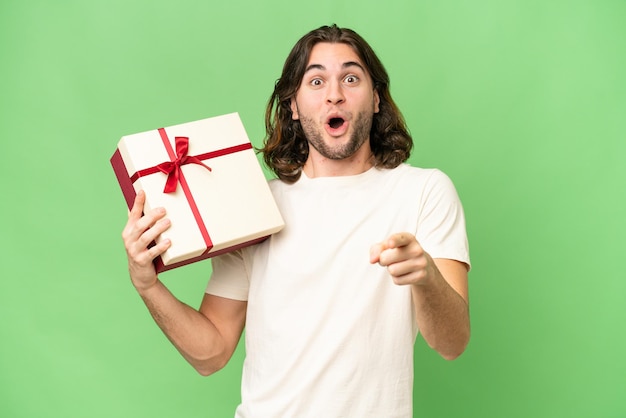 Hombre guapo joven sosteniendo un regalo sobre un fondo aislado sorprendido y apuntando al frente