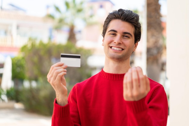 Hombre guapo joven sosteniendo una tarjeta de crédito al aire libre invitando a venir con la mano Feliz de que hayas venido