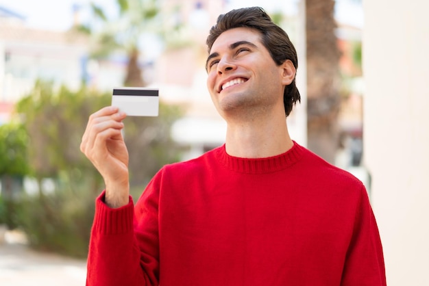 Hombre guapo joven sosteniendo una tarjeta de crédito al aire libre mirando hacia arriba mientras sonríe