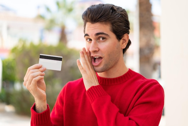 Hombre guapo joven sosteniendo una tarjeta de crédito al aire libre susurrando algo