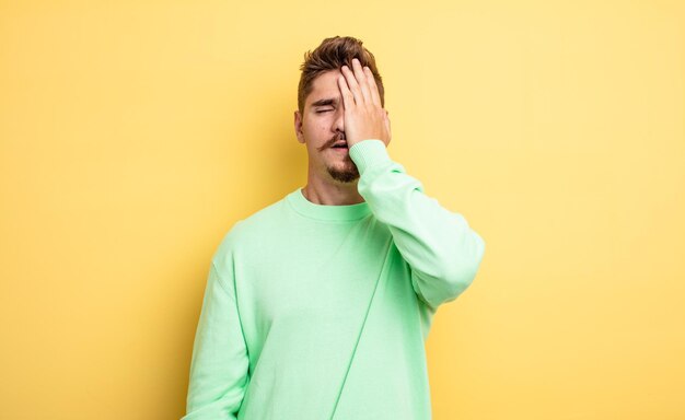 Hombre guapo joven con sueño, aburrido y bostezando, con dolor de cabeza y una mano cubriendo la mitad de la cara. extraño concepto de bigote