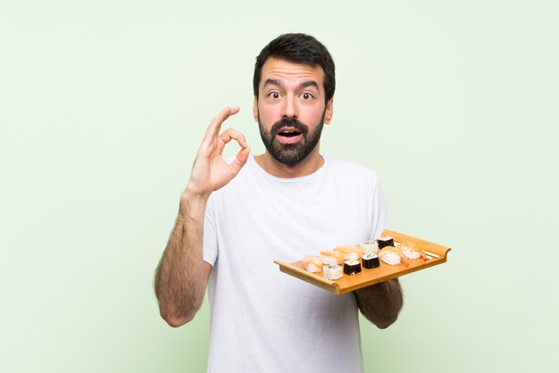 Hombre guapo joven con sushi sobre pared verde aislado sorprendido y mostrando signo ok