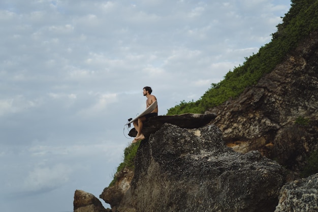 Hombre guapo joven con una tabla de surf en una roca cerca del océano.