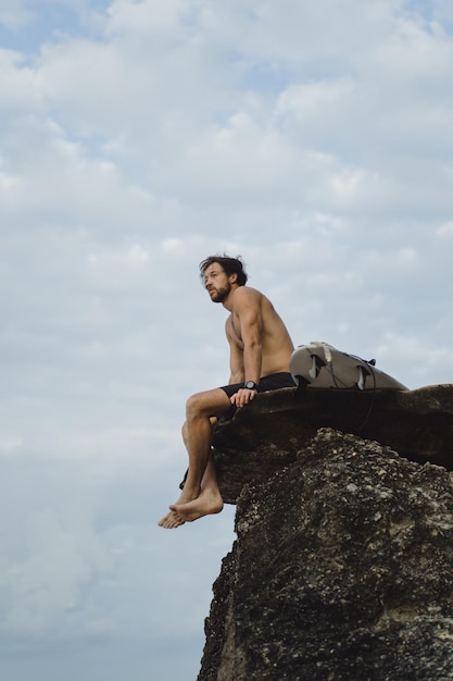 Hombre guapo joven con una tabla de surf en una roca cerca del océano.