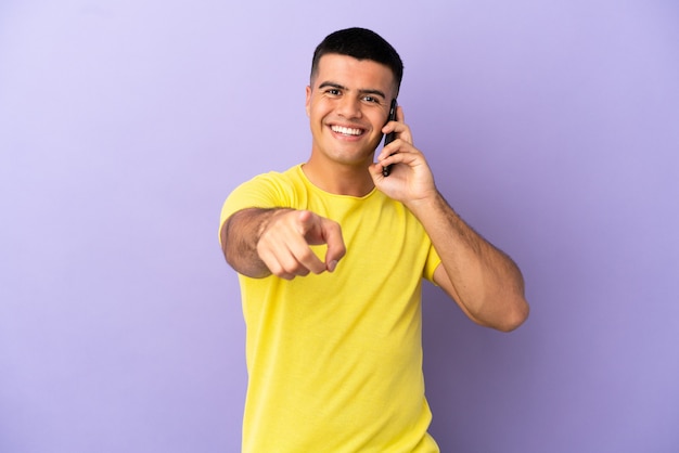 Hombre guapo joven con teléfono móvil sobre fondo púrpura aislado apuntando al frente con expresión feliz