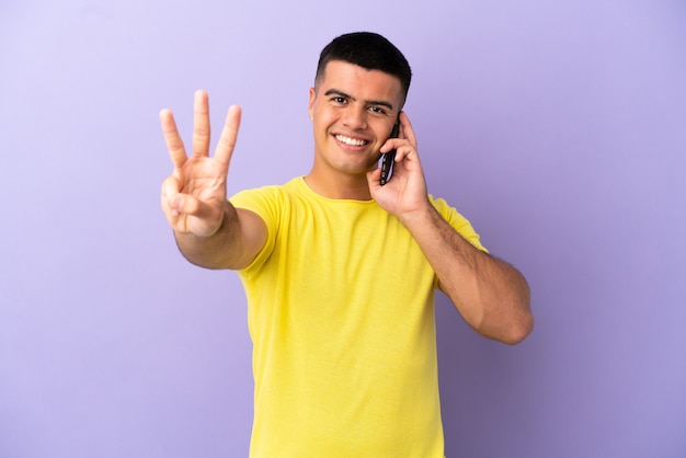 Hombre guapo joven con teléfono móvil sobre fondo púrpura aislado feliz y contando tres con los dedos