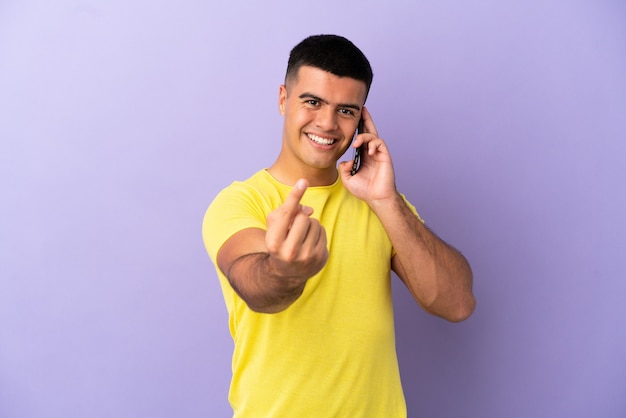 Hombre guapo joven con teléfono móvil sobre fondo púrpura aislado haciendo gesto de venida