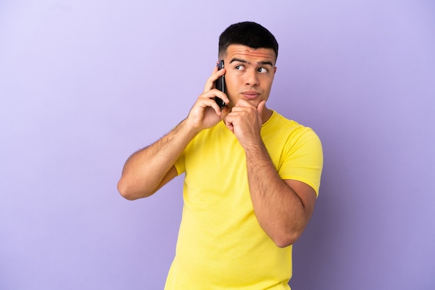Hombre guapo joven con teléfono móvil sobre fondo púrpura aislado y mirando hacia arriba