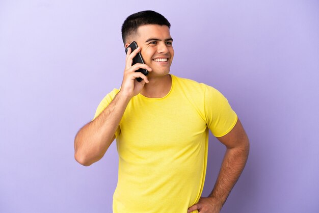 Hombre guapo joven con teléfono móvil sobre fondo púrpura aislado posando con los brazos en la cadera y sonriendo