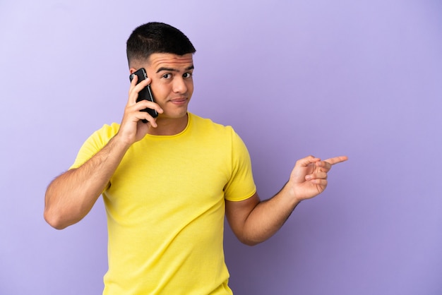 Hombre guapo joven con teléfono móvil sobre fondo púrpura aislado que señala con el dedo hacia el lado