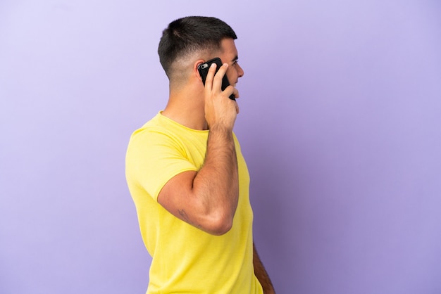 Hombre guapo joven con teléfono móvil sobre fondo púrpura aislado riendo en posición lateral