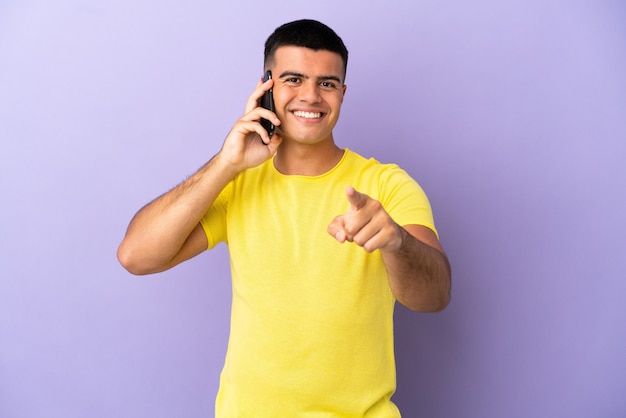 Hombre guapo joven con teléfono móvil sobre fondo púrpura aislado sorprendido y apuntando hacia el frente