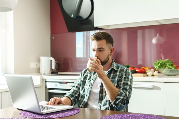 Hombre guapo joven tomando café o té y usando una computadora portátil en la cocina