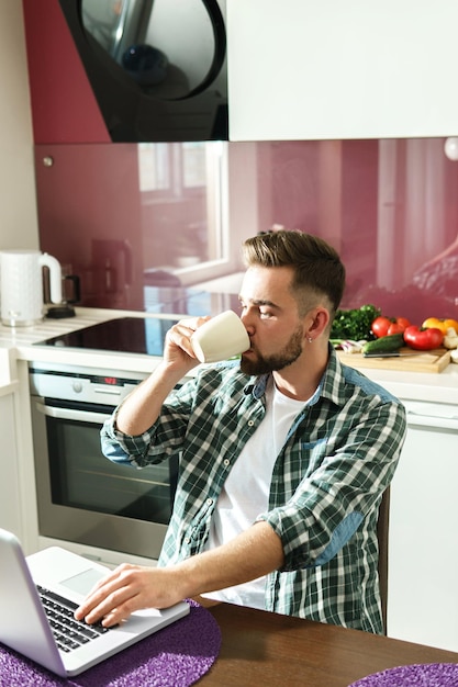 Hombre guapo joven tomando café o té y usando una computadora portátil en la cocina