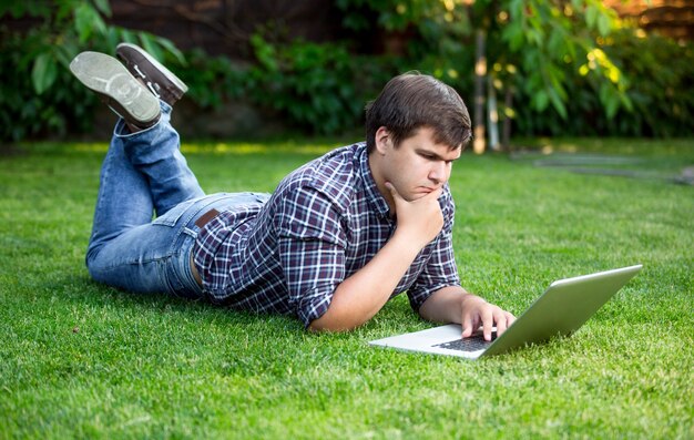 Hombre guapo joven tumbado en la hierba en el parque y usando laptop