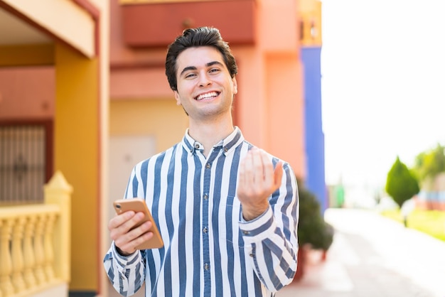 Hombre guapo joven usando teléfono móvil al aire libre invitando a venir con la mano Feliz de que hayas venido