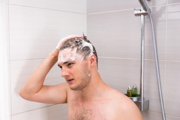 Hombre guapo lavándose el cabello mojado y con espuma en la cabina de ducha en el moderno baño de azulejos