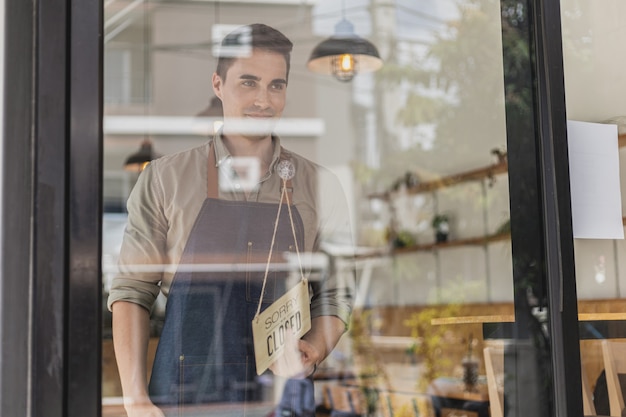 Un hombre guapo con un letrero frente a una tienda, un empleado de un café de pie con un letrero que dice abrir-cerrar, y un letrero que dice "cerrado". Concepto de servicio de comida y bebida de café.
