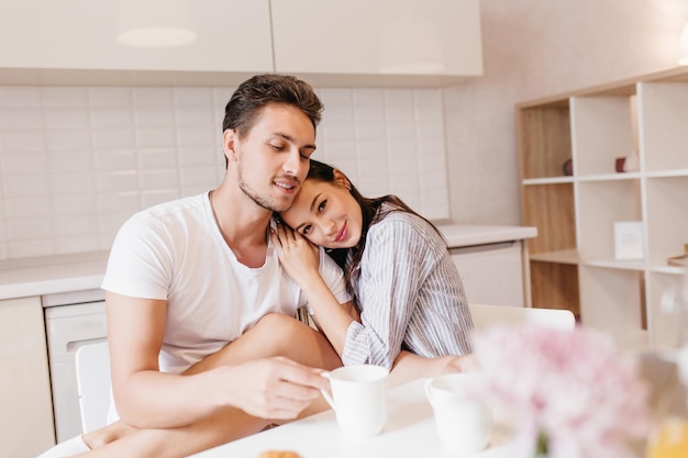 Hombre guapo lleva anillo bebiendo capuchino en la cocina blanca, sentado con su linda novia. Retrato interior de una joven romántica en pijama azul abrazando a su marido mientras disfruta de un café.