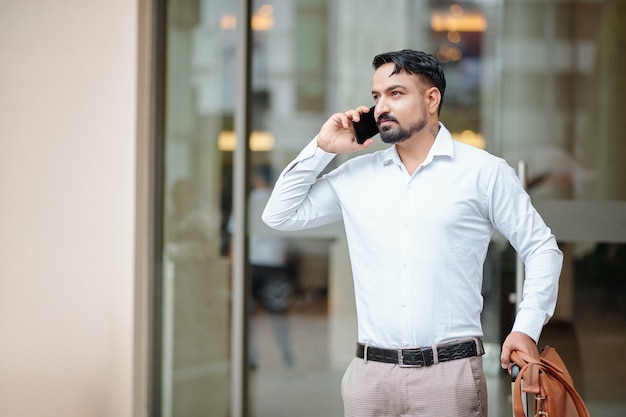 Hombre guapo con maleta y bolso de cuero hablando por teléfono mientras espera un taxi en la entrada del hotel