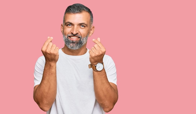 Hombre guapo de mediana edad con camiseta blanca casual haciendo gestos de dinero con las manos pidiendo pago de salario negocio millonario