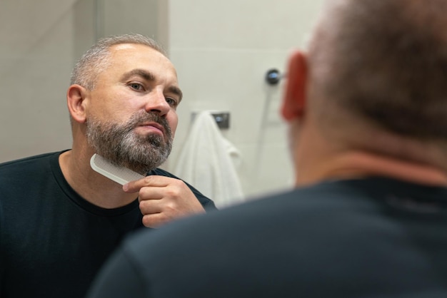 Foto hombre guapo de mediana edad cepillarse la barba por la mañana en el baño mirando en el espejo