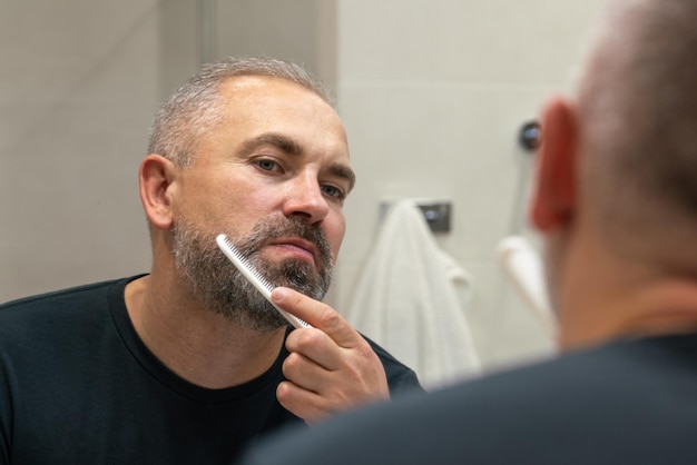 Hombre guapo de mediana edad cepillarse la barba por la mañana en el baño mirando en el espejo