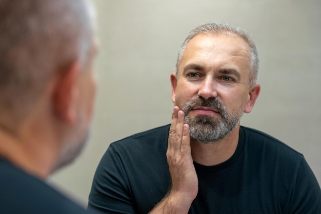 Hombre guapo de mediana edad mirando en el espejo en el baño tocando su barba pensando en cortársela