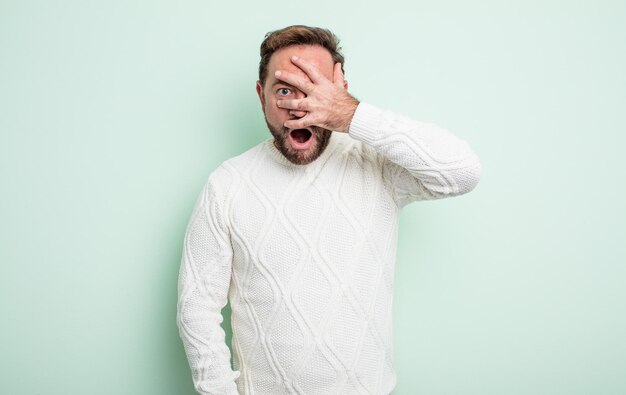 Hombre guapo de mediana edad que parece sorprendido asustado o aterrorizado cubriendo la cara con la mano y mirando entre los dedos