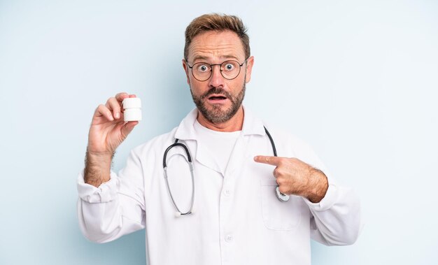 Hombre guapo de mediana edad que se siente feliz y apunta a sí mismo con un entusiasmo. médico con botella de pastillas