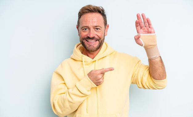 Hombre guapo de mediana edad sonriendo alegremente, sintiéndose feliz y apuntando hacia un lado. concepto de mano rota