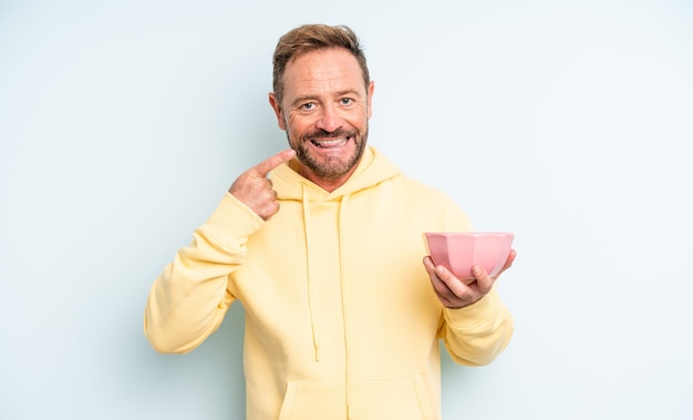 Hombre guapo de mediana edad sonriendo con confianza apuntando a su propia amplia sonrisa. concepto de cuenco vacío