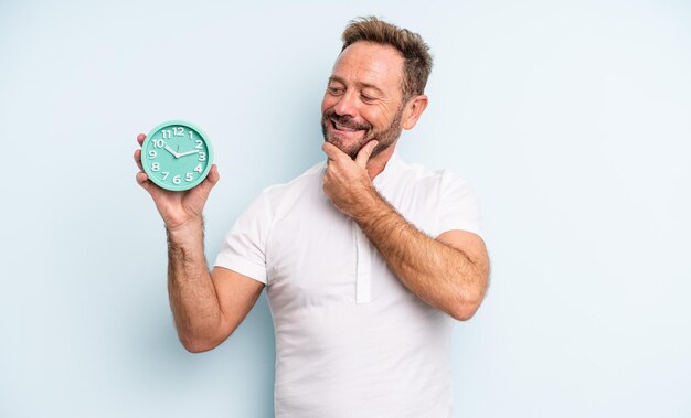 Hombre guapo de mediana edad sonriendo con una expresión feliz y segura con la mano en la barbilla. concepto de reloj despertador