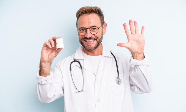 Hombre guapo de mediana edad sonriendo y mirando amigable, mostrando el número cinco. médico con botella de pastillas