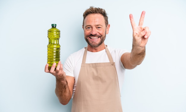 Hombre guapo de mediana edad sonriendo y mirando feliz, gesticulando victoria o paz. concepto de aceite de oliva