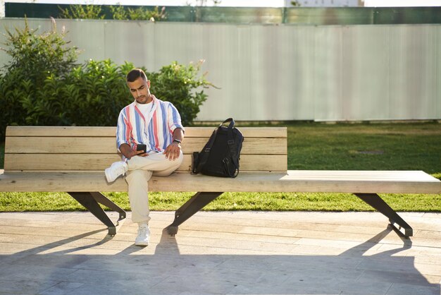Hombre guapo del medio oriente usando teléfono móvil, comunicación en línea sentado en el banco