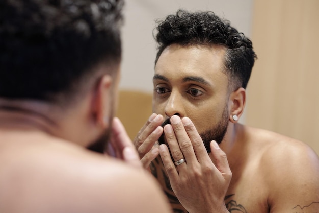 Hombre guapo mirando en el espejo después de tomar la ducha matutina y aplicar crema hidratante