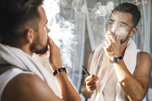 Hombre guapo mirando en el espejo, fumando un cigarrillo y afeitándose la barba con una navaja de afeitar