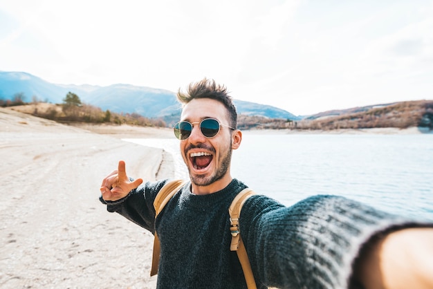 Hombre guapo con mochila tomando selfie excursionista joven al aire libre viajar en las montañas sonriendo a la cámara