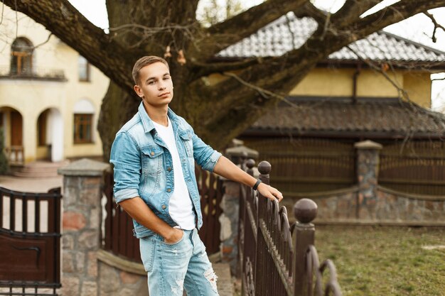 Hombre guapo de moda en una chaqueta de mezclilla con una camiseta blanca y jeans rotos cerca de la valla