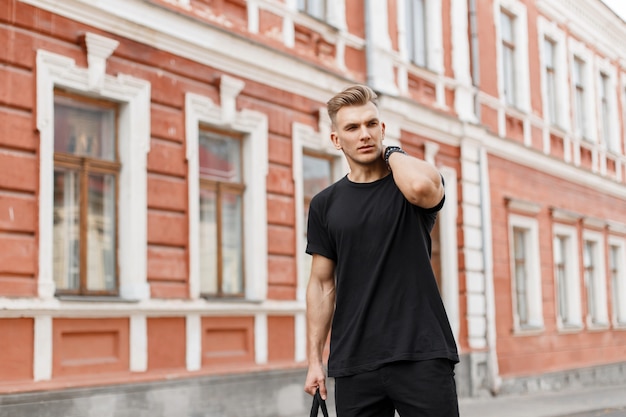 Hombre guapo de moda joven modelo americano con peinado en camiseta negra con bolso negro caminando por la calle en la ciudad